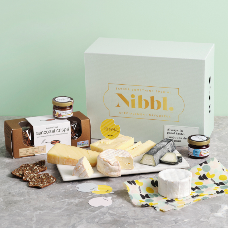 A Nibbl cheese box with various cheeses and crackers spread out over a board 